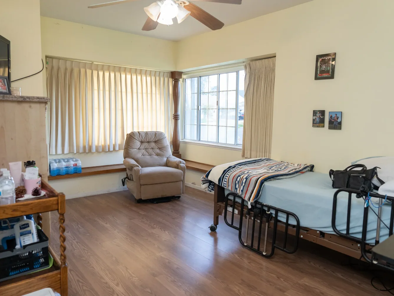 Spacious accessible bedroom featuring hospital bed with safety rails, comfortable recliner, and organized medical supplies in a bright, home-like setting.